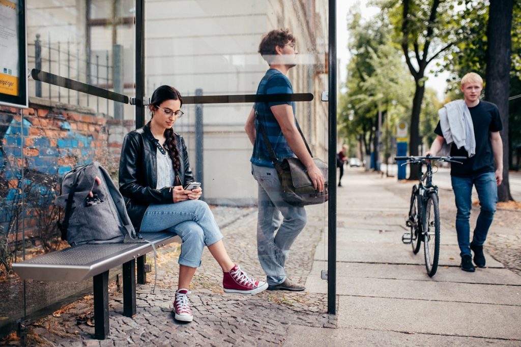 Eine junge Frau sitzt an einer Haltestelle und sieht auf ihr Mobiltelefon. Daneben steht ein Junger Mann, an das Haltestellenhäuschen gelehnt. Ein anderer junger Mann schiebt sein Fahrrad an der Haltestelle vorbei. 
